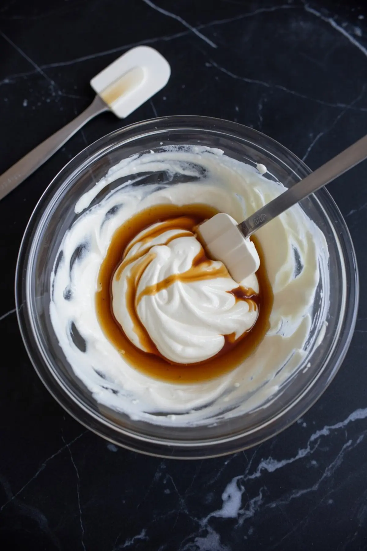 Bowl of whipped cream mixed with vanilla extract and a spatula resting inside, placed on a dark marbled surface, showing the base preparation step for no-churn ice cream.