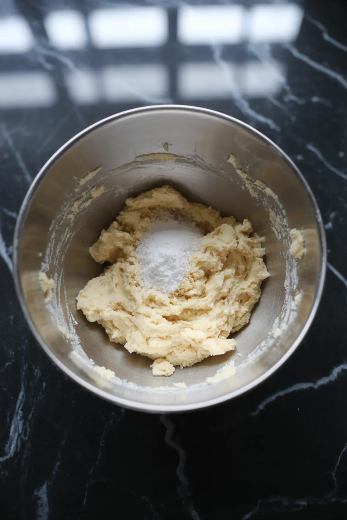A stainless steel mixing bowl filled with creamed dough and a mound of white sugar and salt on top, resting on a dark marble countertop. This is an early stage in preparing the tart crust for mini pecan pies.