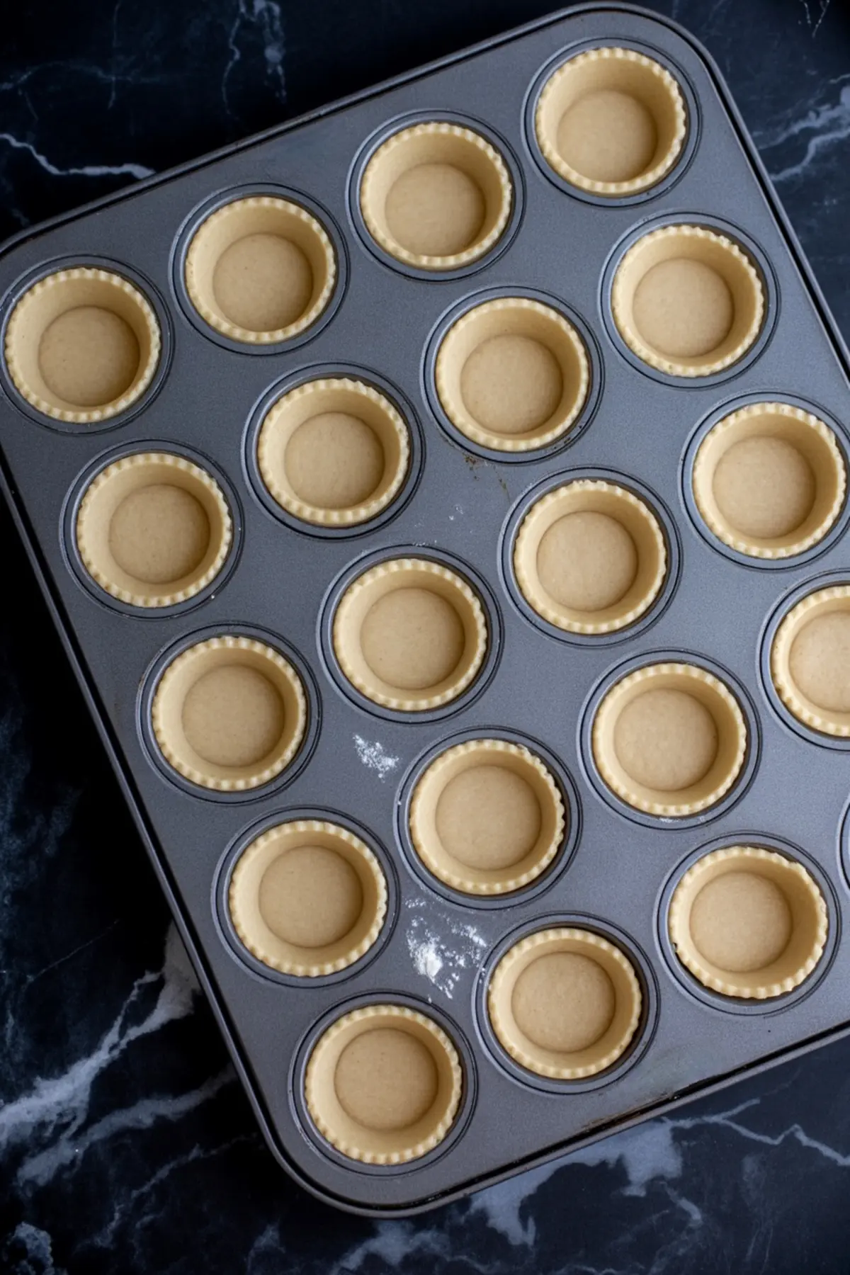 A muffin pan lined with evenly shaped tart dough shells ready to be filled. Each cavity holds an unbaked tart crust with fluted edges, placed on a black marble background.