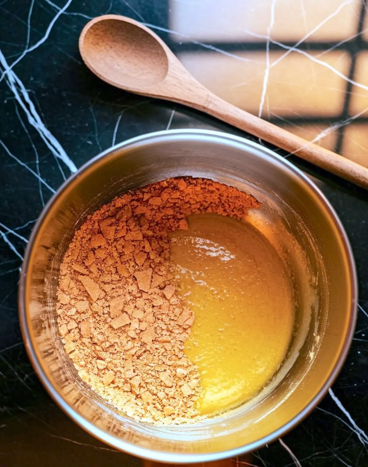 Crushed graham crackers and melted butter being mixed in a stainless steel bowl with a wooden spoon nearby. This step prepares the base for an American cheesecake.
