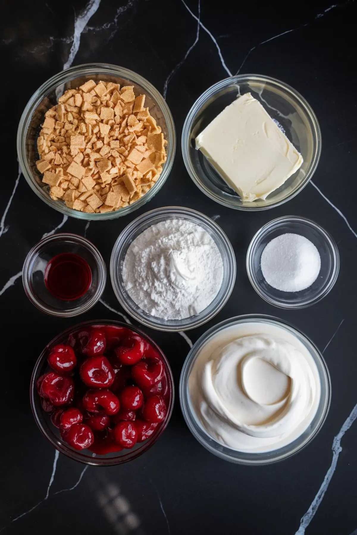 Overhead view of no-bake cherry cheesecake ingredients including crushed vanilla wafers, butter, powdered sugar, granulated sugar, vanilla extract, canned cherries, and whipped cream cheese, all arranged in glass bowls on a black marble countertop.
