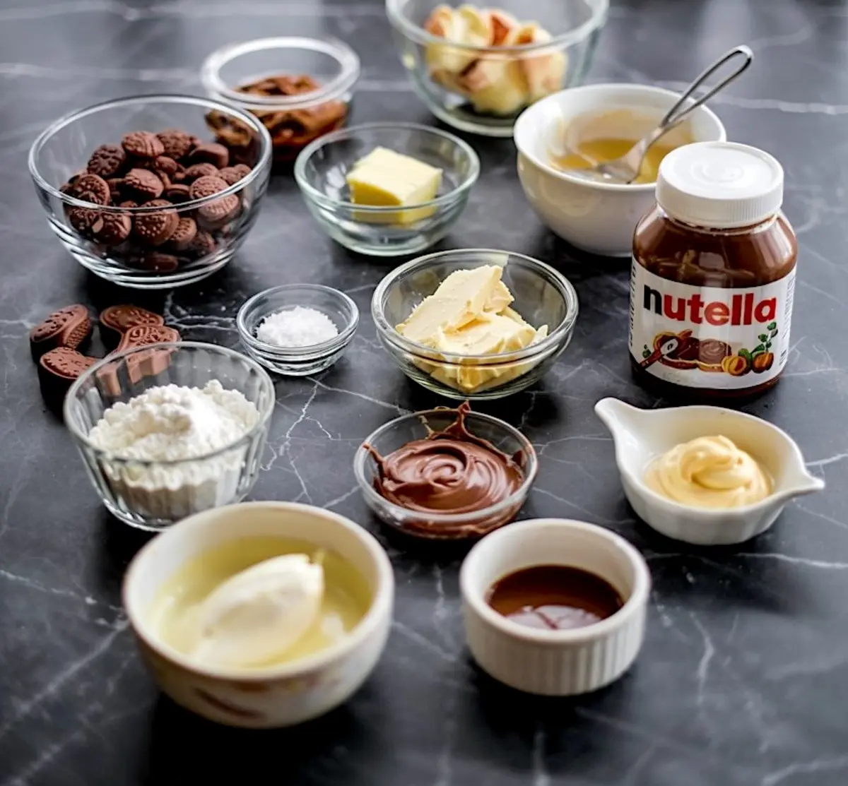 Flat lay of Nutella cheesecake ingredients including Nutella jar, cream cheese, cocoa powder, chocolate cookies, and butter, staged on black marble for Nutella dessert recipe inspiration.