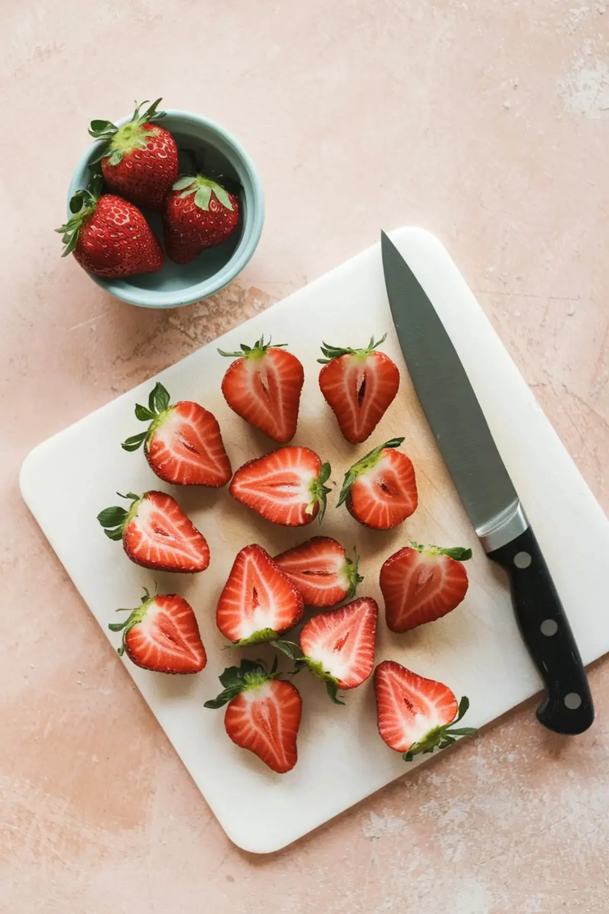 Fresh strawberries cut in half and arranged on a white cutting board with a knife beside a small bowl of whole strawberries, all placed on a light pink surface.
