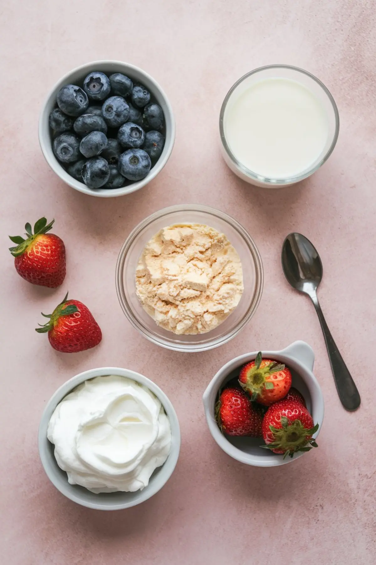 Ingredients for a pudding pie arranged on a pink surface, including fresh strawberries, blueberries, whipped topping, vanilla pudding mix, and a glass of milk in individual bowls and cups.