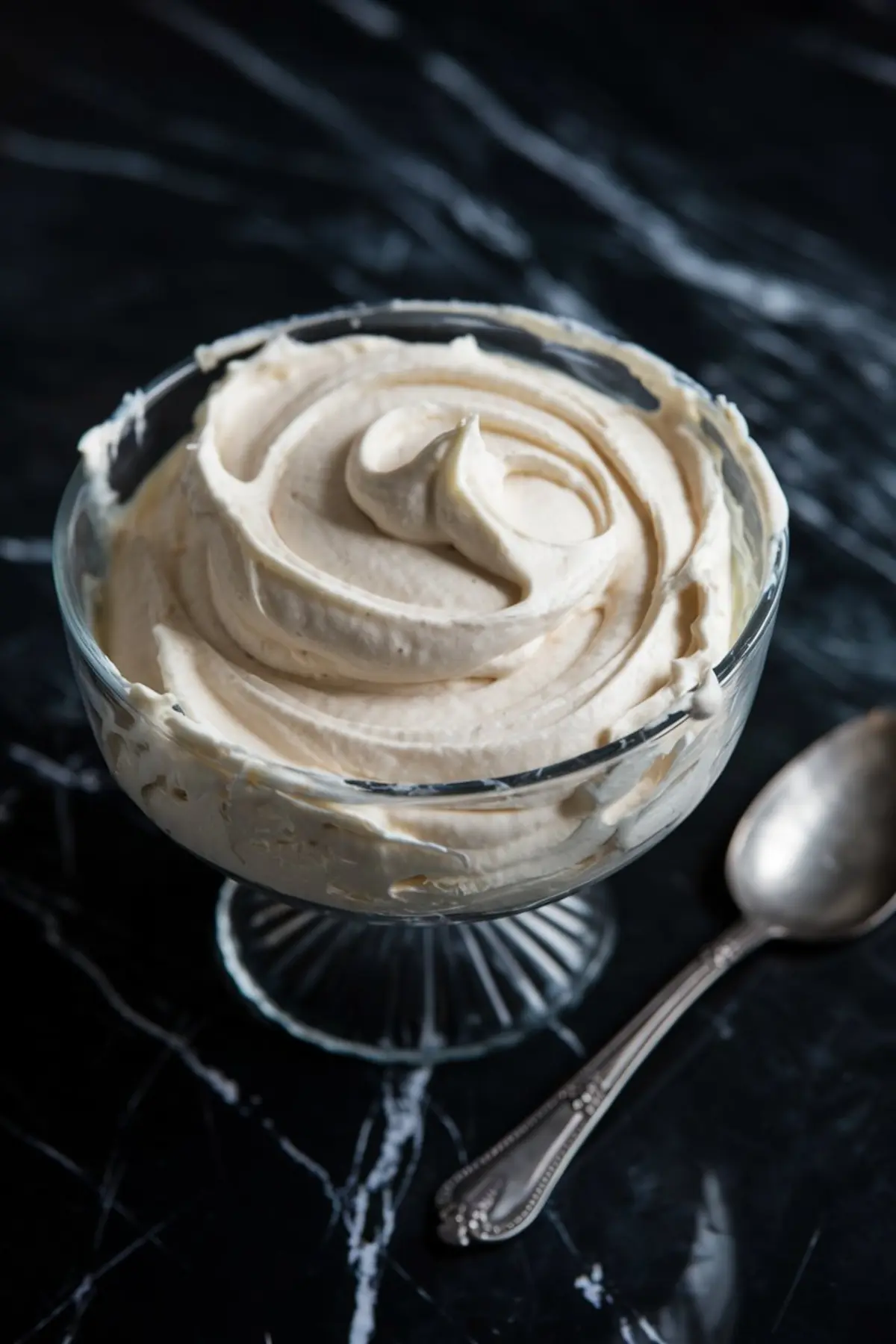 Close-up of smooth vanilla buttercream in a clear glass dessert dish, placed on a dark marble counter with a silver spoon nearby, capturing the texture and richness of the macaron filling.