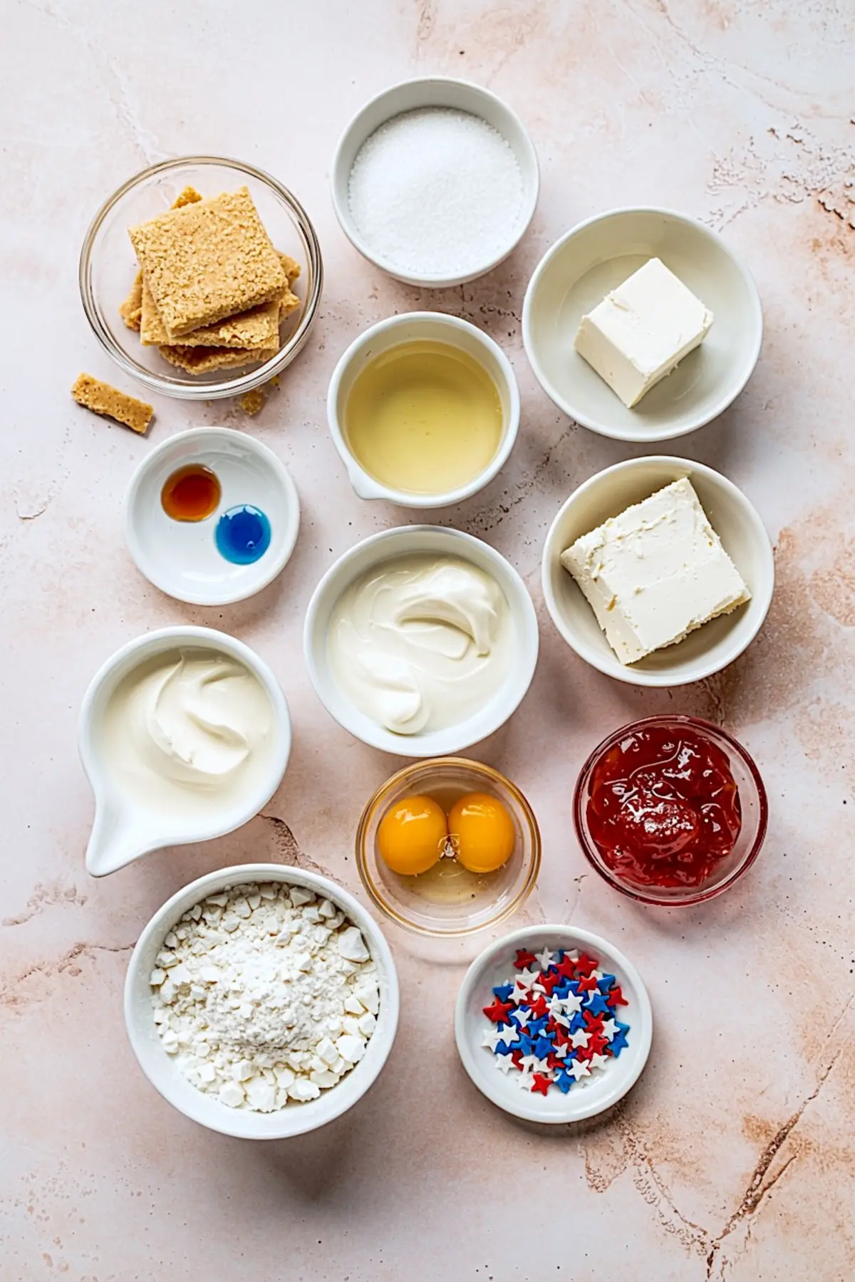 Flat lay of cheesecake ingredients including graham crackers, sugar, cream cheese, sour cream, eggs, vanilla, red jam, blue coloring, and patriotic star sprinkles.