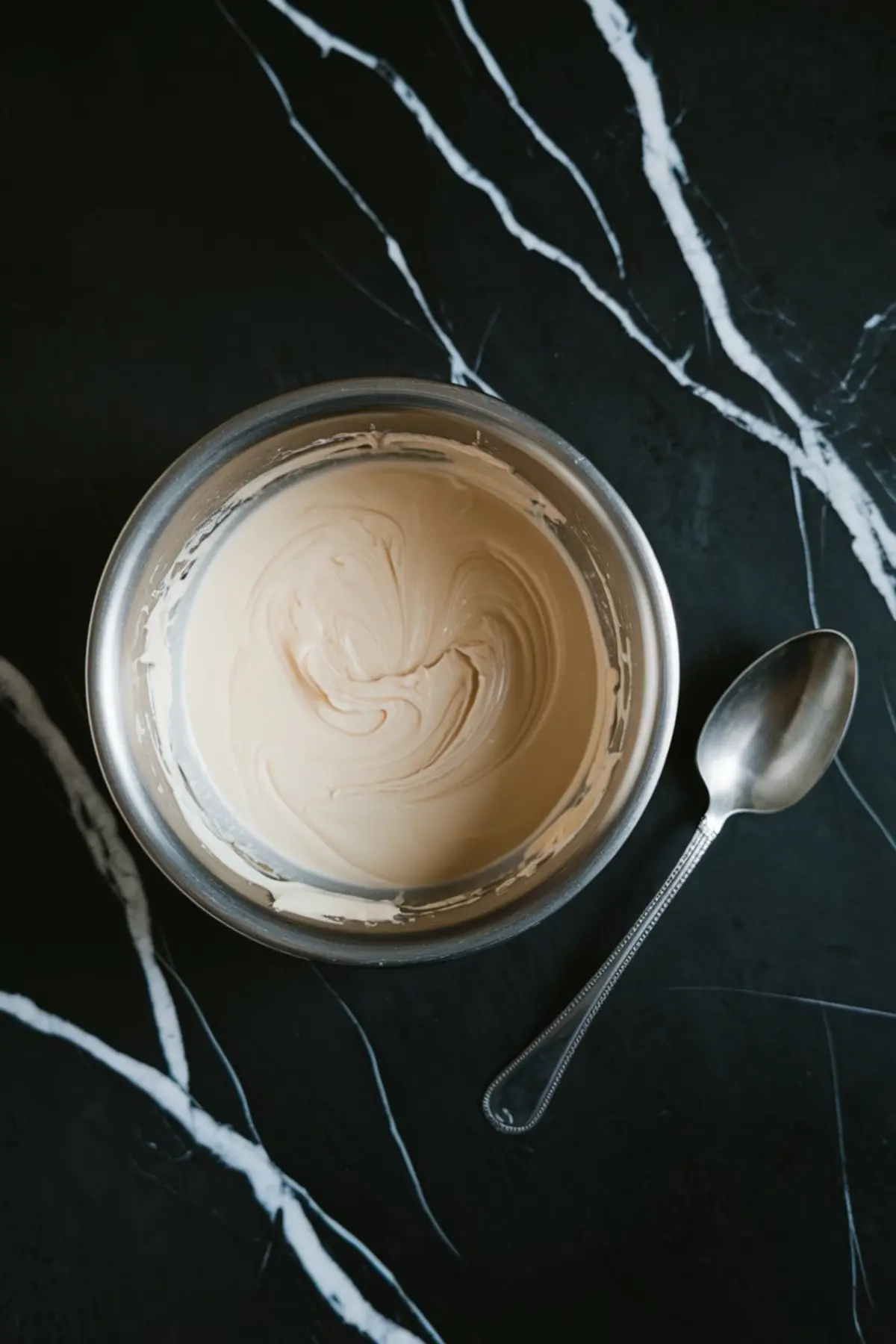 Stainless steel bowl filled with smooth, swirled white chocolate, placed next to a silver spoon on a black marble surface, ready for dessert dipping.