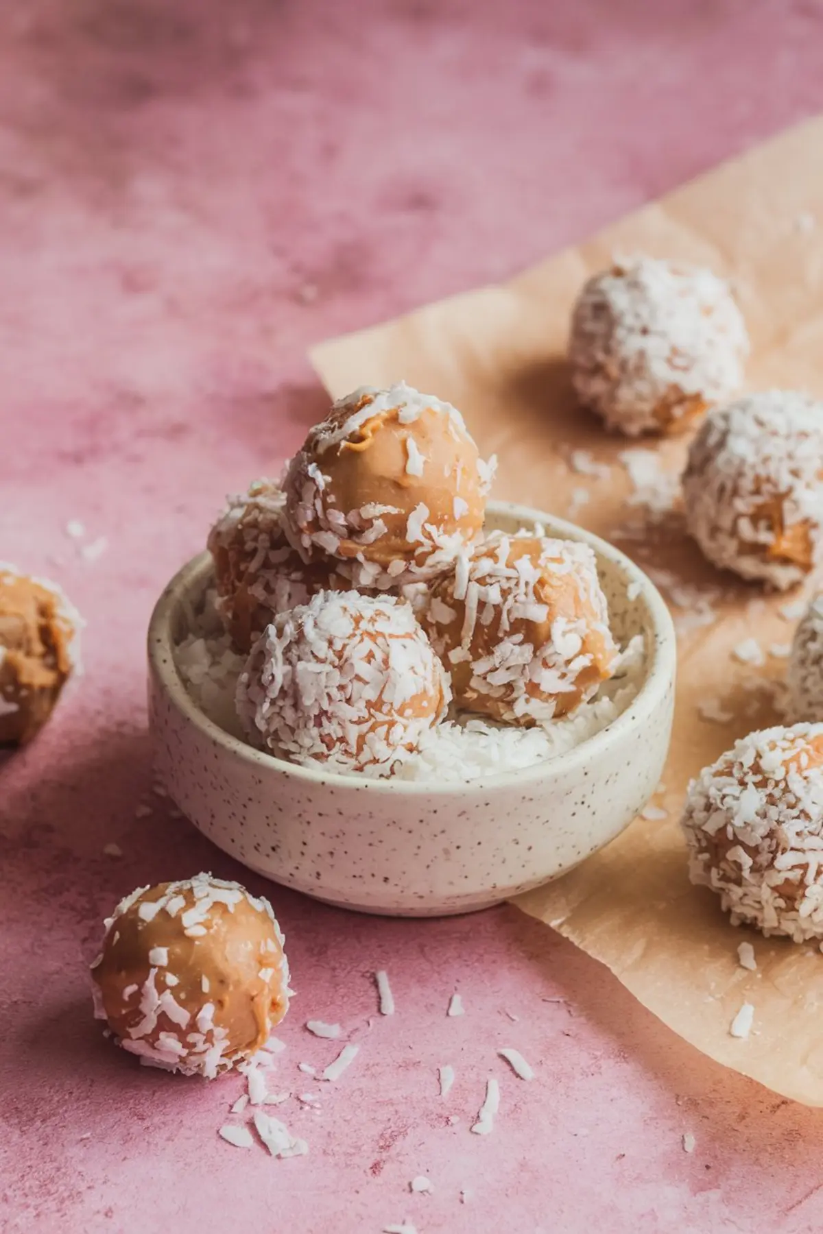 Speckled ceramic bowl filled with no-bake peanut butter cookies coated in shredded coconut, with cookies scattered on a pink surface and parchment paper.
