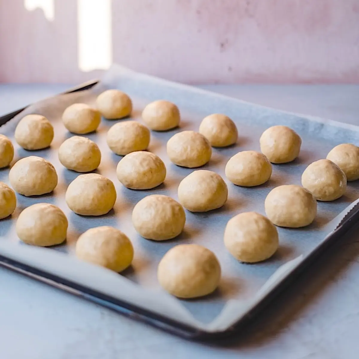 Raw cookie dough balls evenly spaced on a parchment-lined baking sheet, ready for baking with soft natural lighting and a pink background.
