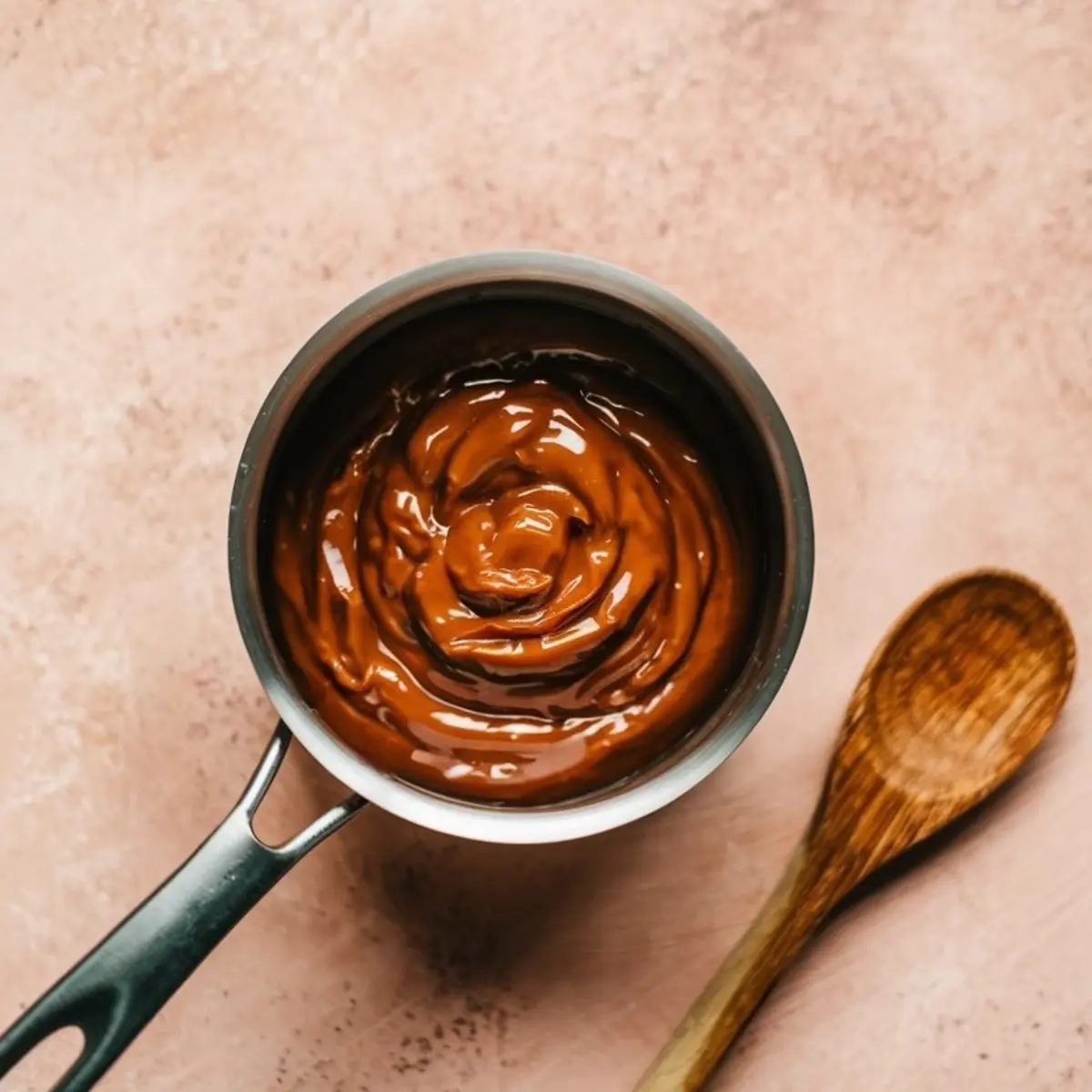 Small saucepan filled with smooth, swirled brown caramel filling next to a wooden spoon on a peach-toned background.