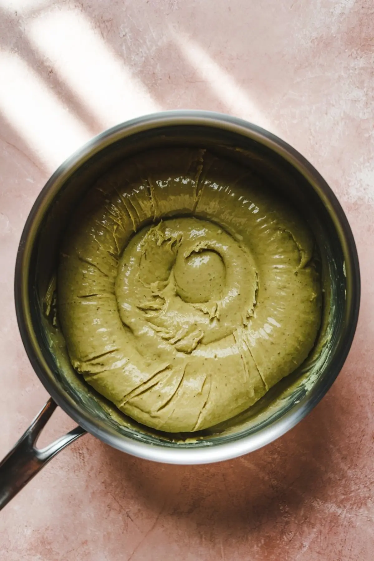 Overhead shot of thick pistachio brigadeiro dough swirled in a saucepan on a pink surface, showcasing its smooth, creamy texture.