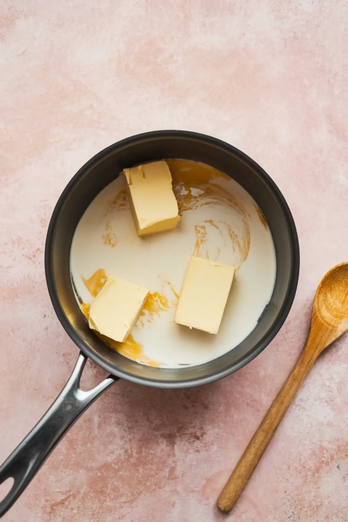 Overhead image of a saucepan with three blocks of butter melting into sweetened condensed milk on a pink countertop, with a wooden spoon placed beside it, showing the initial stage of making pistachio brigadeiro.