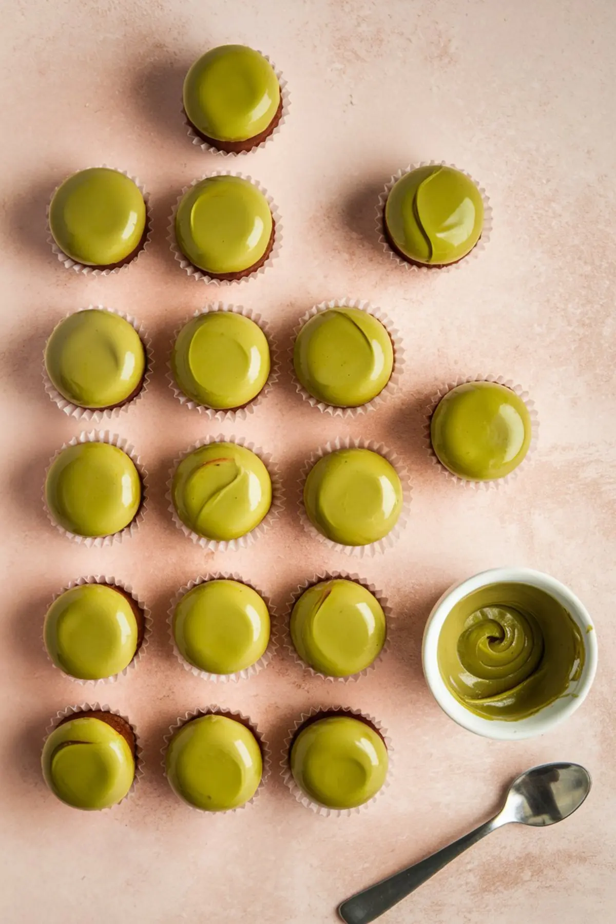 Flat lay of glossy pistachio-glazed mini cupcakes arranged in a geometric grid on a blush pink background, with a small bowl of creamy pistachio ganache and a spoon beside them.