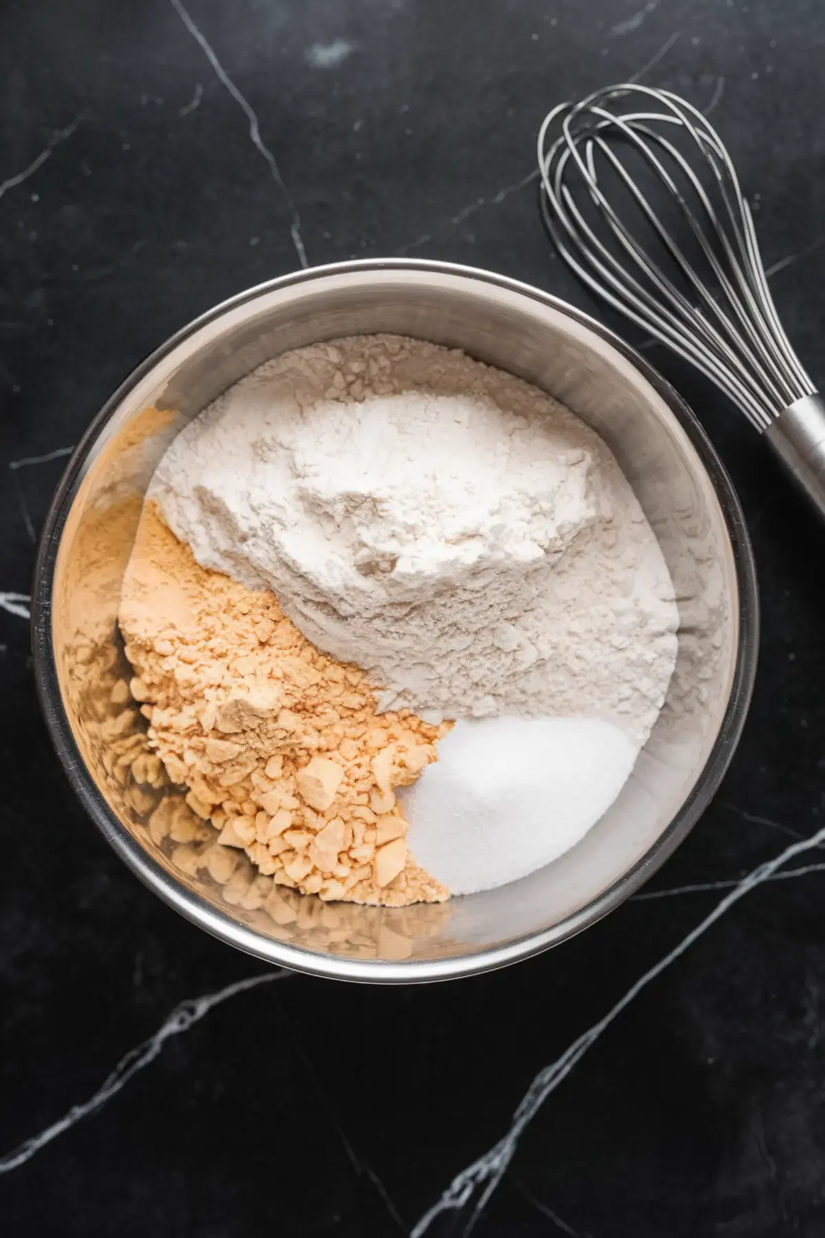 Stainless steel mixing bowl containing flour, ground nuts, and sugar, displayed in sections before mixing, next to a metal whisk on a dark marble surface.