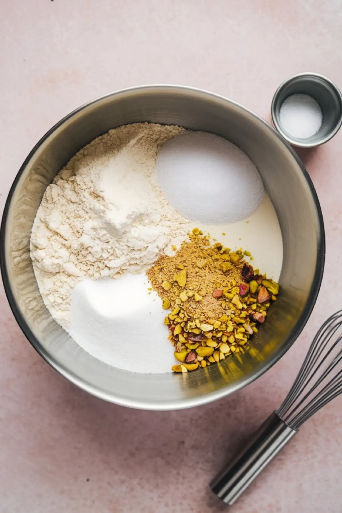 Dry ingredients in a mixing bowl for pistachio muffins including flour, sugar, ground pistachios, and baking powder, with a whisk and salt dish on a pink countertop.
