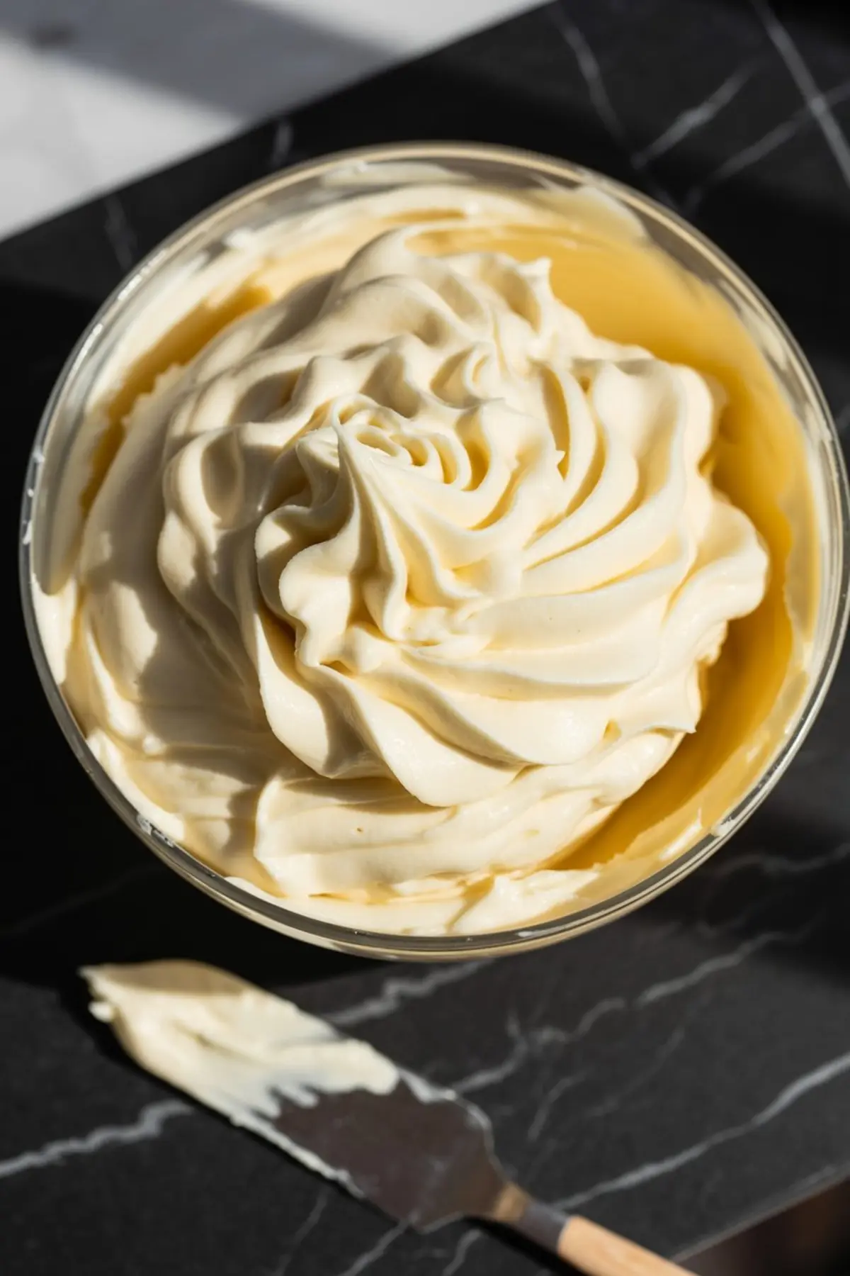 Swirled vanilla pudding cream filling in a glass bowl with a metal spatula, ready for sandwich cookie assembly, set on a dark marble countertop.