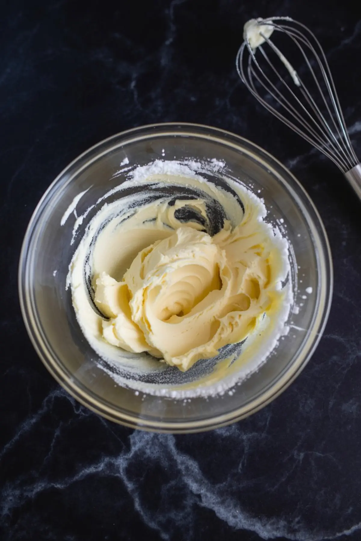 Buttercream frosting whipped in a glass bowl with powdered sugar visible, with a metal whisk resting on a black marble surface.