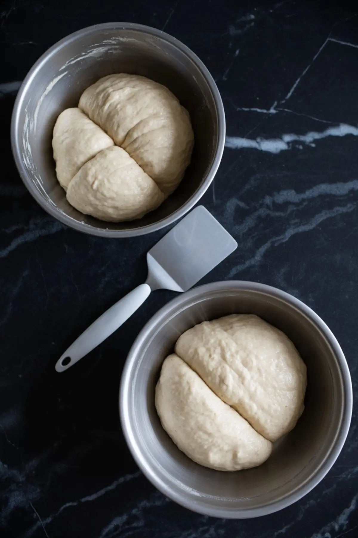 Two bowls of risen dough divided into sections, placed in metal mixing bowls with a white dough scraper on a dark marble countertop.