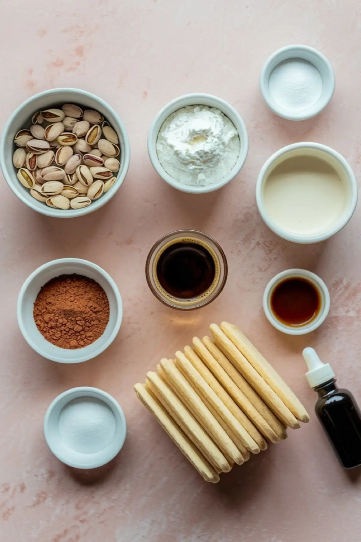 Flat lay of pistachio tiramisu ingredients including pistachios, mascarpone, cocoa powder, ladyfinger cookies, espresso, vanilla extract, sugar, heavy cream, and pistachio paste on a pink backdrop.

