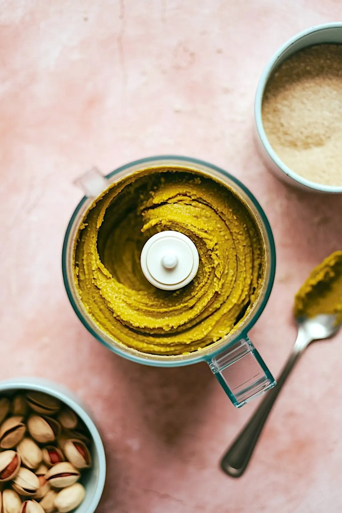 Top view of freshly blended pistachio paste in a food processor surrounded by raw pistachios and brown sugar on a pink countertop.
