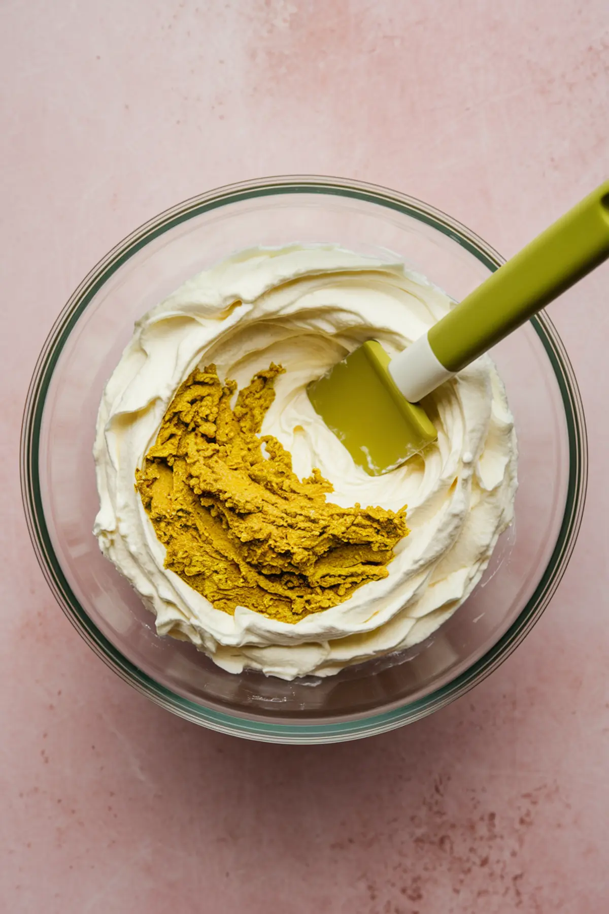 Close-up of pistachio paste being folded into whipped mascarpone cream in a glass mixing bowl with a green spatula on a light pink surface.
