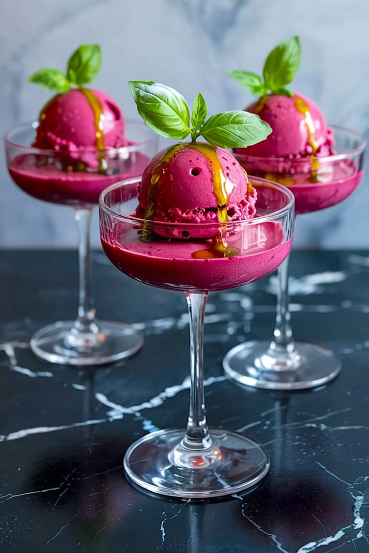 Three glass dessert cups filled with deep purple plum sorbet scoops, topped with basil oil drizzle and fresh basil leaves, arranged on a dark marble table.