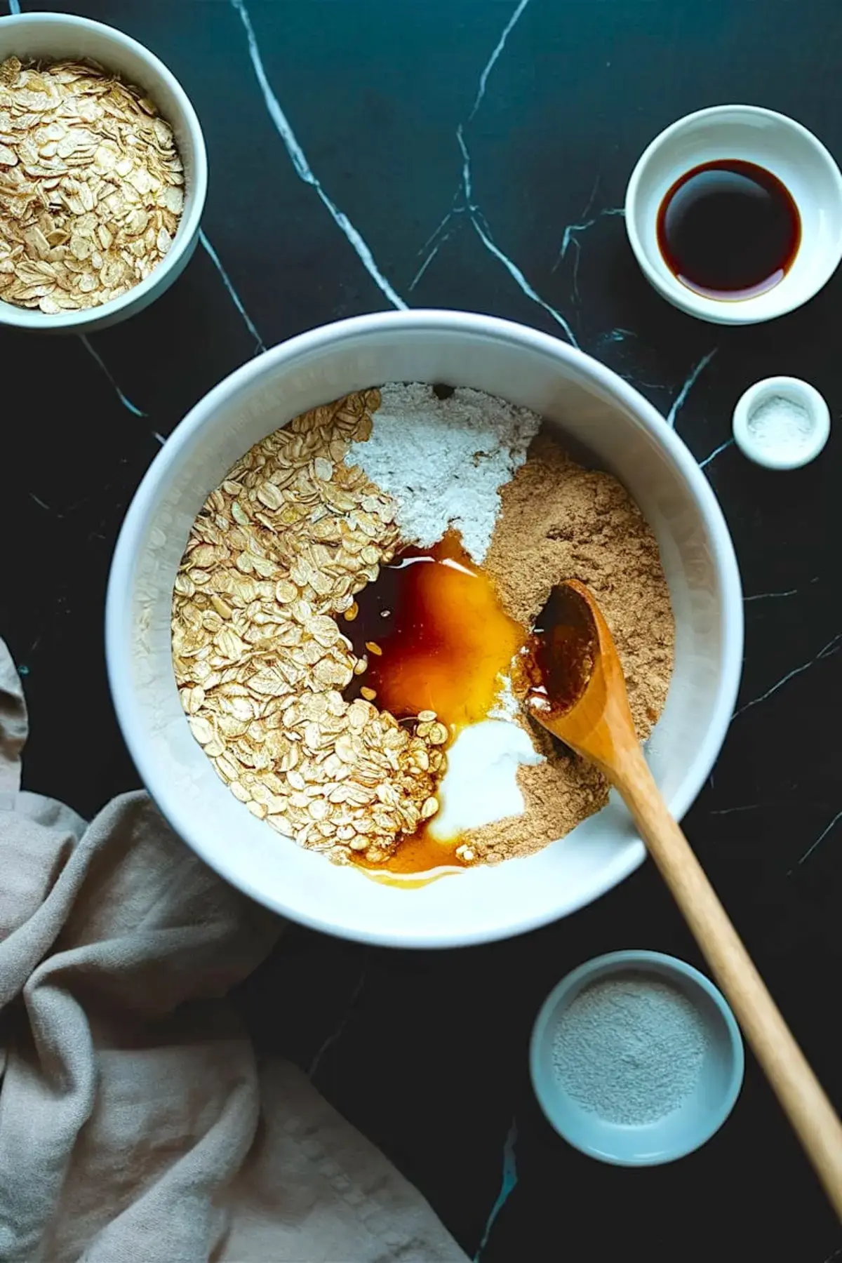 Overhead view of a mixing bowl with rolled oats, brown sugar, protein powder, baking soda, vanilla extract, and syrup on a dark marble surface, showing ingredients for a high-protein dessert base.