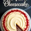 Overhead view of a whole red velvet cheesecake with cream cheese frosting and red cake crumbs around the edge, with one slice removed, displayed on a white marble cake stand with “Red Velvet Cheesecake” text above.