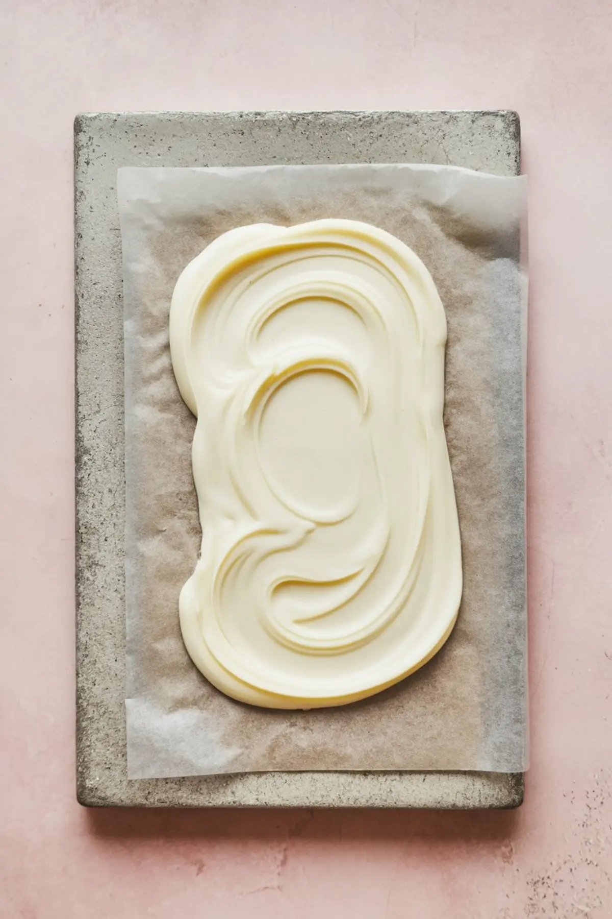 Tray lined with parchment paper holding freshly spread melted white chocolate, swirled in smooth circular motions. The baking tray rests on a soft pink background, emphasizing a minimal and clean preparation stage for chocolate bark recipes.