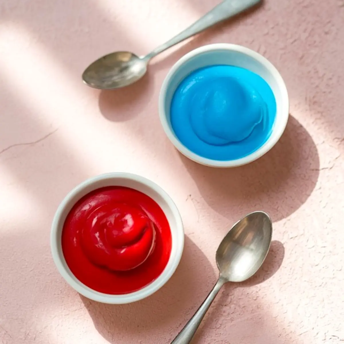 Overhead shot of two small bowls of vibrant red and bright blue candy melts or food coloring, placed on a pink textured surface with metal spoons nearby. The image emphasizes baking ingredients used for colorful dessert decorating or themed chocolate bark preparation.