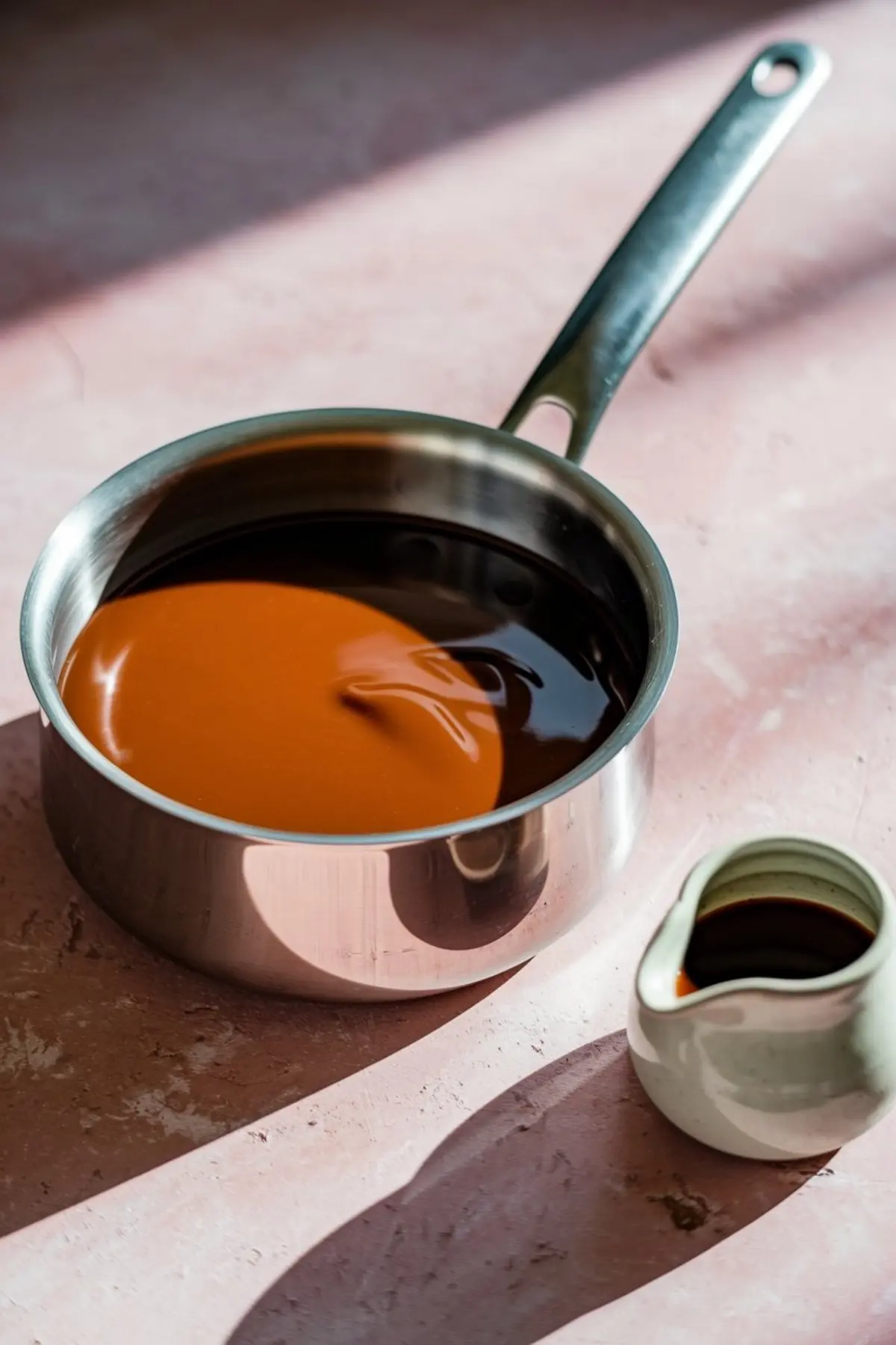 Saucepan filled with homemade toffee sauce, partially swirled with dark syrup, next to a small cream jug on a pink kitchen counter.