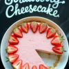 Overhead photo of a pink strawberry cheesecake with one slice removed, garnished with halved fresh strawberries and placed over a black cloth with “Strawberry Cheesecake” written in white script.