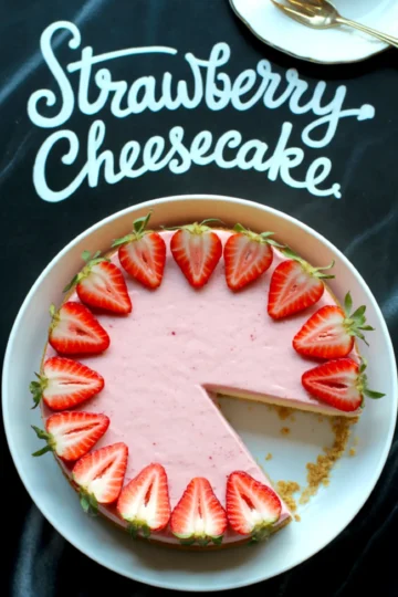 Overhead photo of a pink strawberry cheesecake with one slice removed, garnished with halved fresh strawberries and placed over a black cloth with “Strawberry Cheesecake” written in white script.