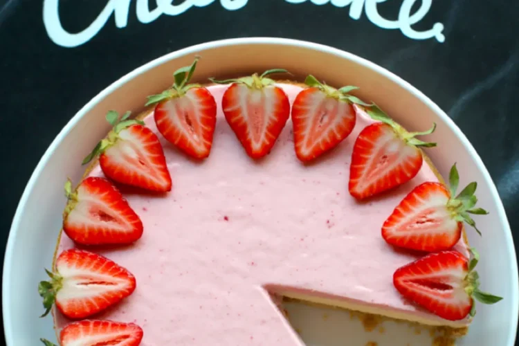 Overhead photo of a pink strawberry cheesecake with one slice removed, garnished with halved fresh strawberries and placed over a black cloth with “Strawberry Cheesecake” written in white script.