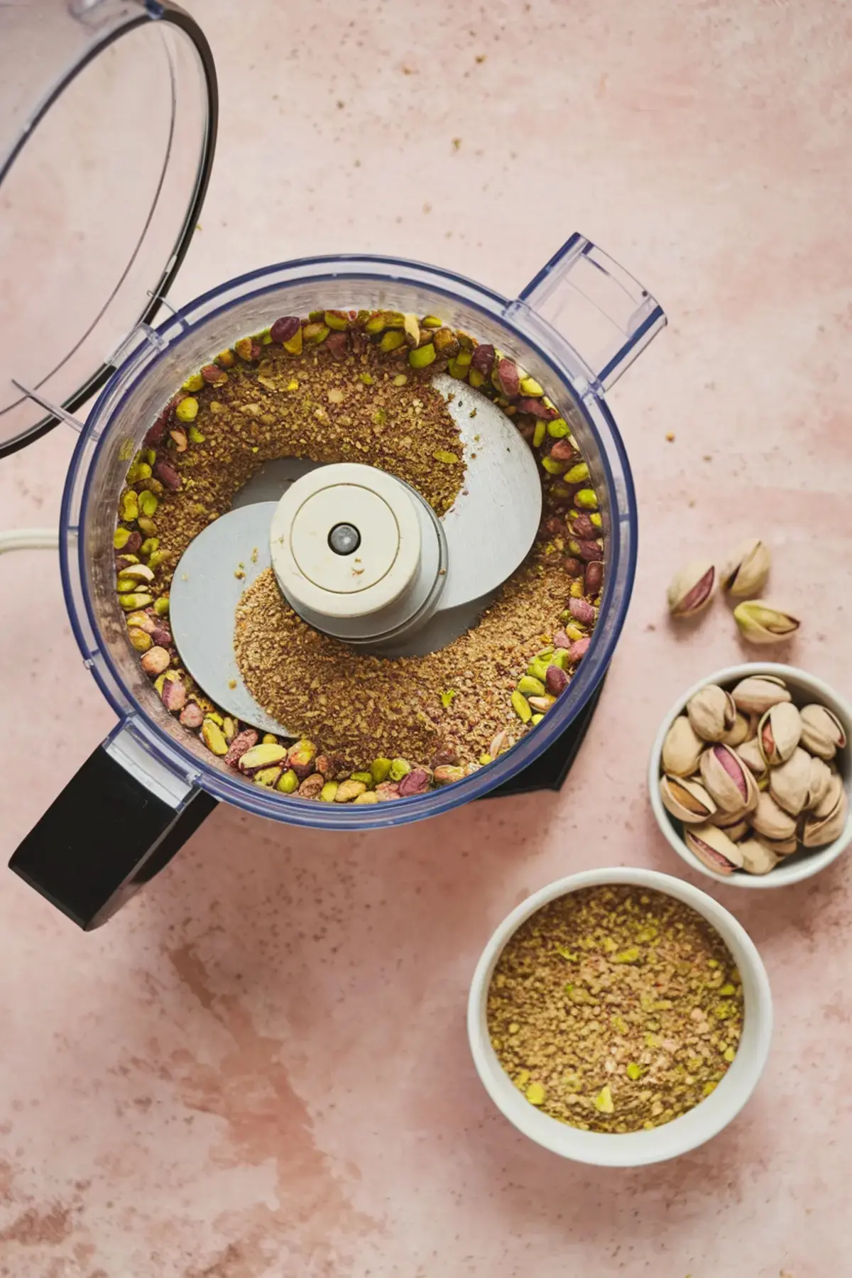 Pistachios being ground in a food processor with whole and crushed nuts in white bowls on a pink surface.
