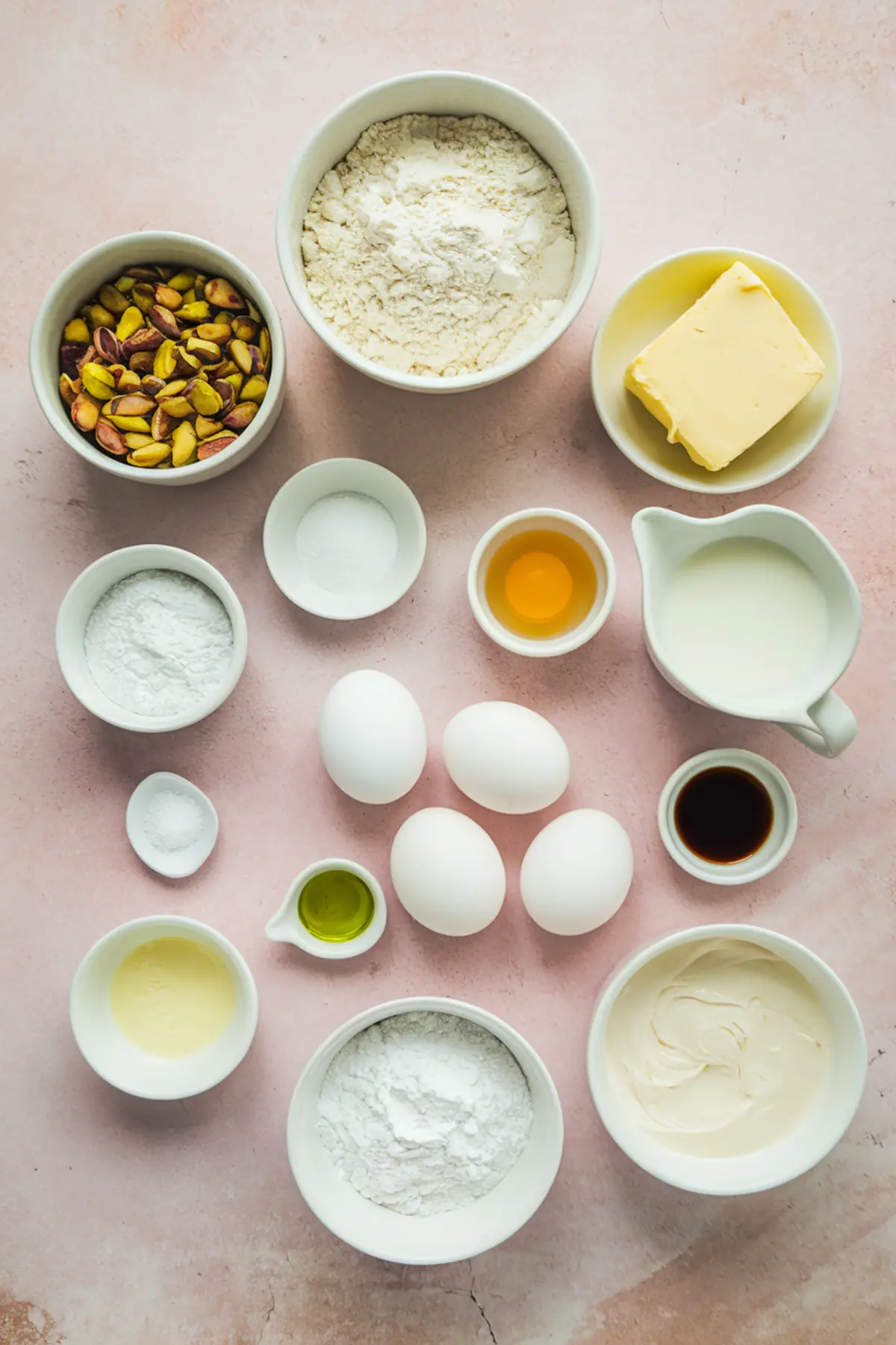 Flat lay of baking ingredients including pistachios, flour, eggs, butter, and milk arranged in small bowls.