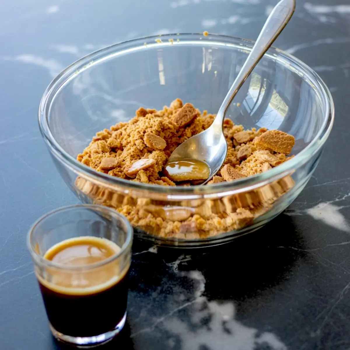 Crushed speculoos cookies in a glass bowl with a spoon and a shot of espresso on a black marble countertop, ready for tiramisu cheesecake crust preparation.
