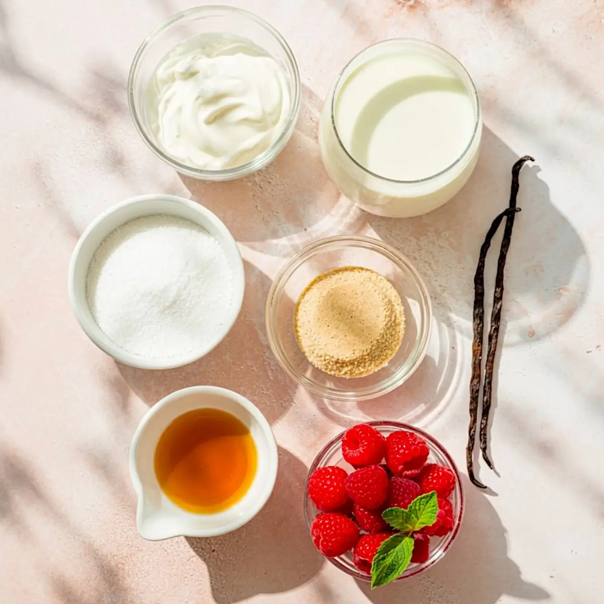Flat lay of vanilla panna cotta ingredients including sugar, gelatin, cream, yogurt, vanilla extract, fresh raspberries with mint, and whole vanilla beans on a pale pink background.
