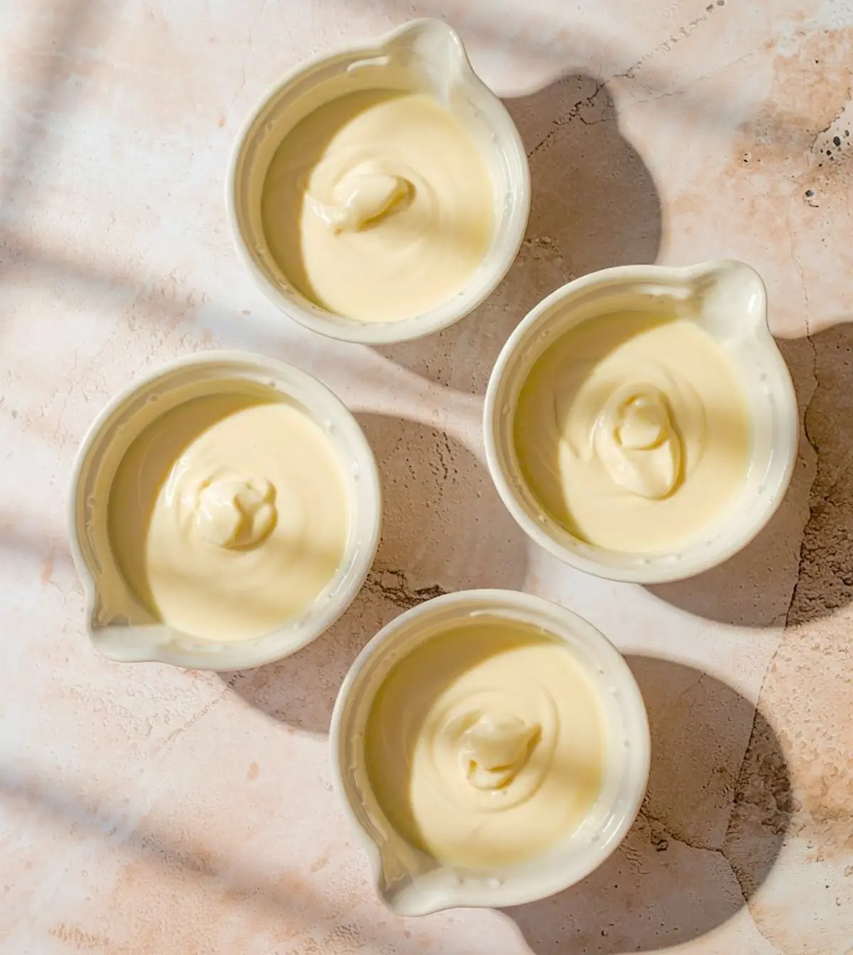 Four white ramekins filled with freshly poured vanilla panna cotta mixture, set on a sunlit pink stone surface ready to chill.
