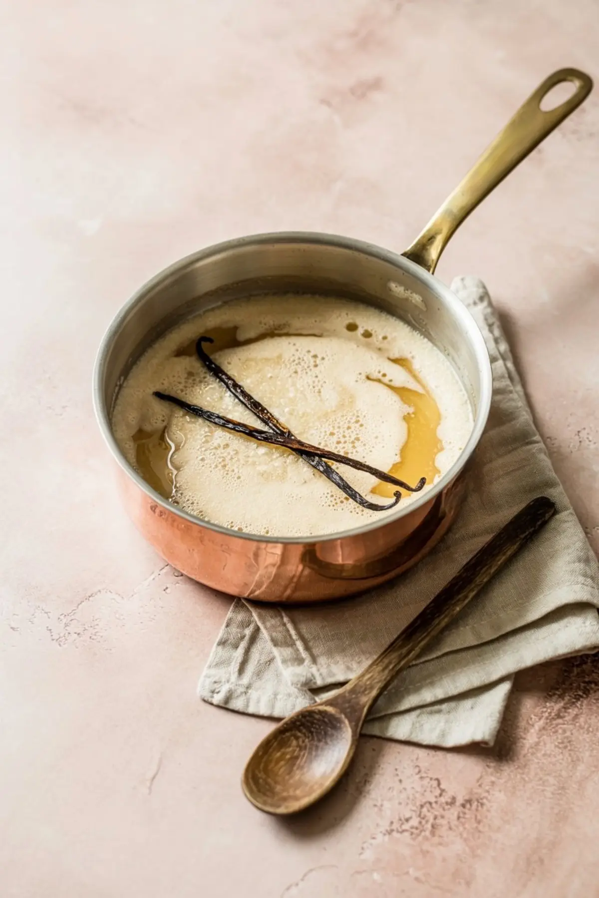 Saucepan of vanilla cream mixture with whole vanilla beans, lightly foaming and infused, placed on a beige napkin with a wooden spoon on a pink countertop.

