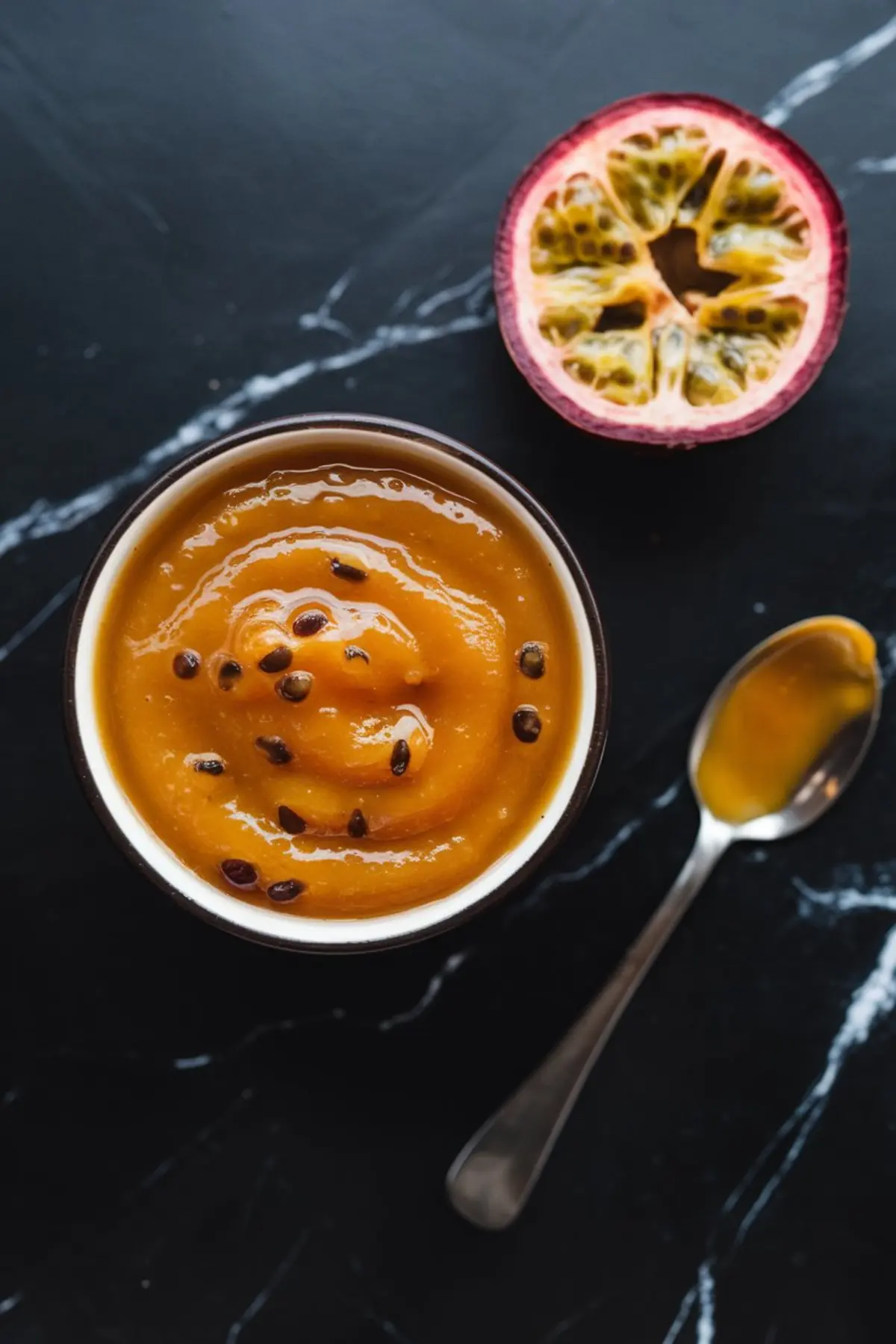 Bowl of thick passion fruit curd with seeds, placed next to a halved passion fruit and spoon on a black surface, ready for topping or plating.
