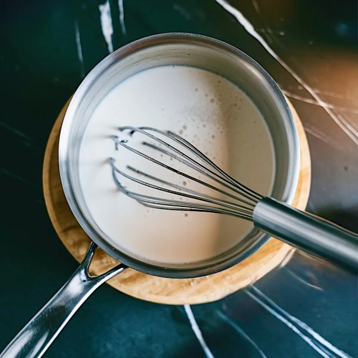 Overhead view of a saucepan with white liquid being whisked, set on a wooden trivet over a dark countertop, capturing a step in vegan panna cotta preparation.

