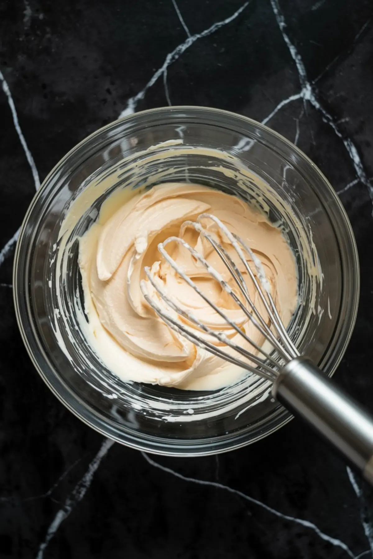 Glass bowl with smooth cream cheese cheesecake batter being mixed using a metal whisk on a black marble countertop.