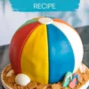 Round cake decorated to look like a colorful beach ball with red, yellow, blue, white, green, and orange fondant panels, placed on a cookie crumb base with edible seashells, graham crackers, and flip-flop decorations.