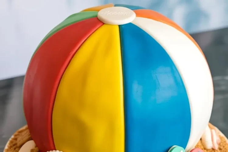 Round cake decorated to look like a colorful beach ball with red, yellow, blue, white, green, and orange fondant panels, placed on a cookie crumb base with edible seashells, graham crackers, and flip-flop decorations.
