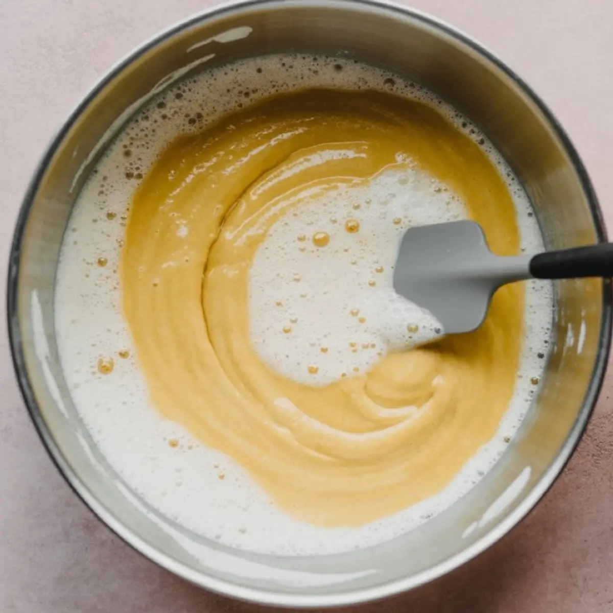 Mixing yellow chiffon cake batter with fluffy egg whites in a metal bowl using a silicone spatula on a pastel pink surface.