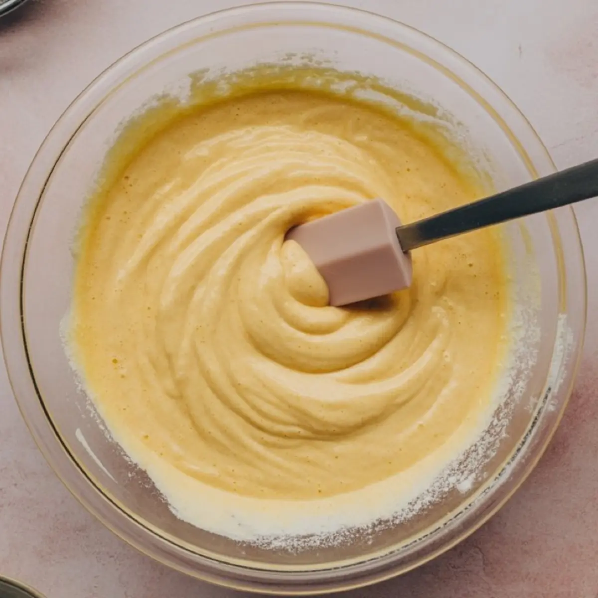 Smooth yellow chiffon cake batter being stirred in a clear glass bowl with a pink spatula on a soft pink background.