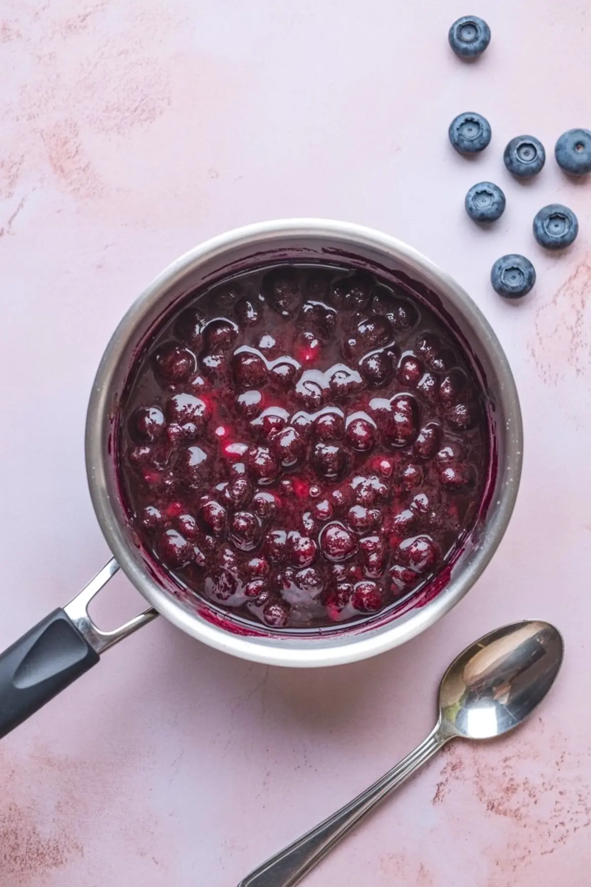 Saucepan filled with glossy homemade blueberry compote, surrounded by fresh blueberries and a spoon on a pink background.
