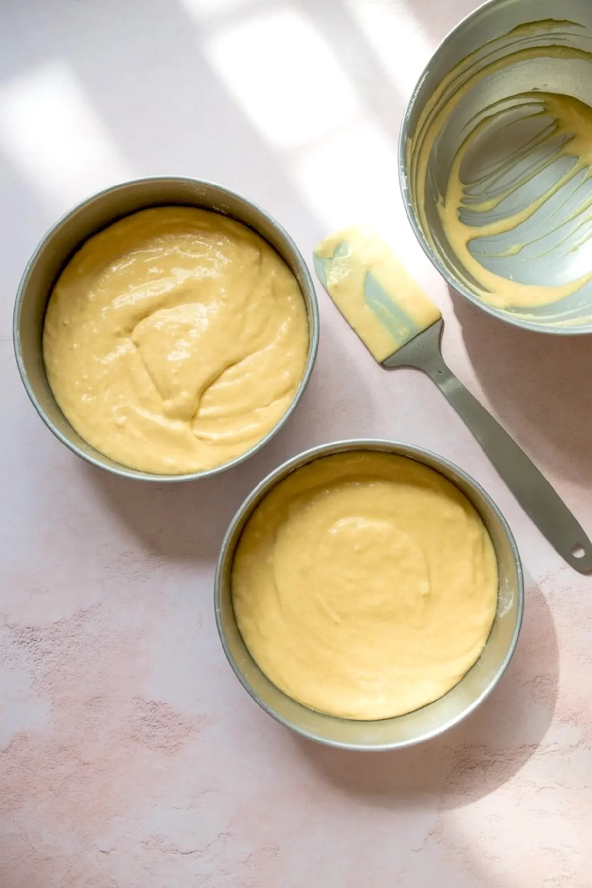 Unbaked chiffon cake batter spread evenly in two round cake pans with an empty bowl and a spatula beside them, ready for baking.
