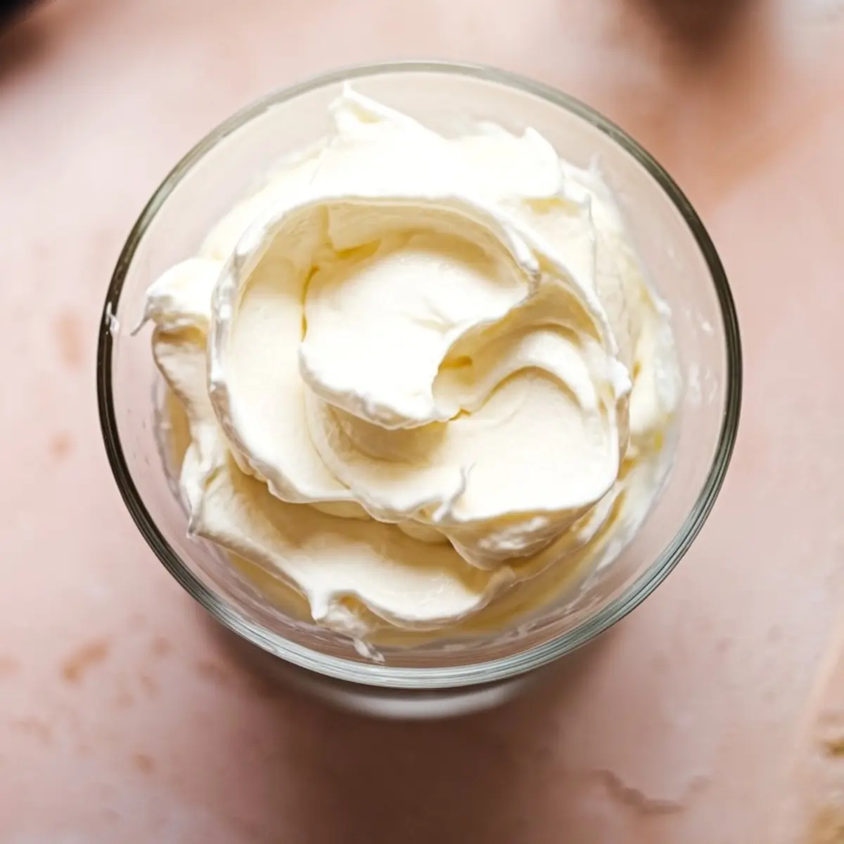 Fluffy whipped cream in a glass bowl with soft peaks and a creamy texture, set on a pale pink background.
